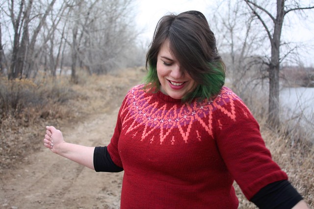 Photo of a plus-sized woman modeling a handknit sweater in red, coral, lilac, and white. 