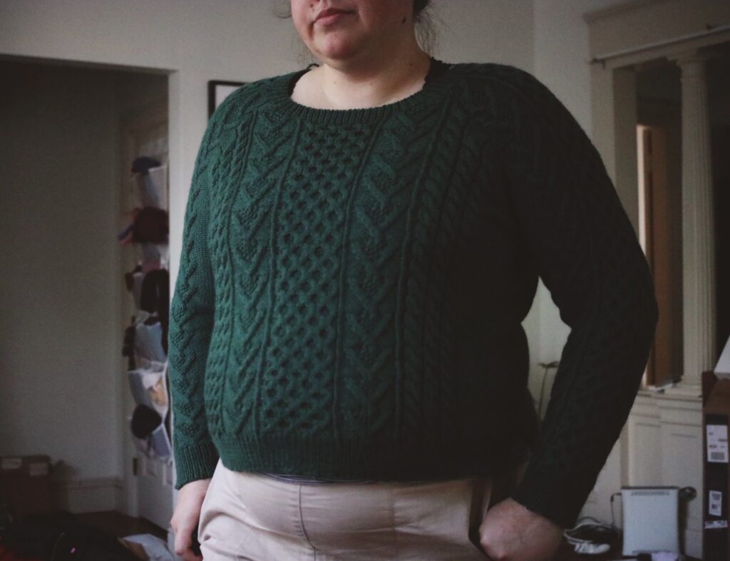 Photo of a plus-sized woman wearing a green cabled sweater and khaki shorts. The pattern is the Bronwyn sweater designed by Melissa Wehrle.