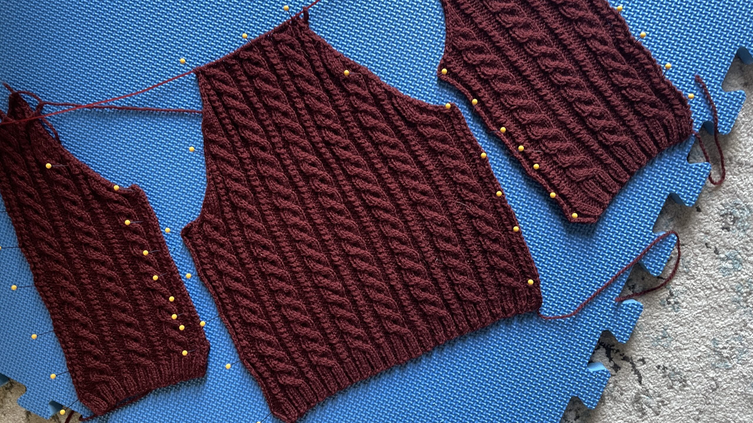 Photo of a small cabled cardigan in progress, with the back and both fronts blocking on the mat.